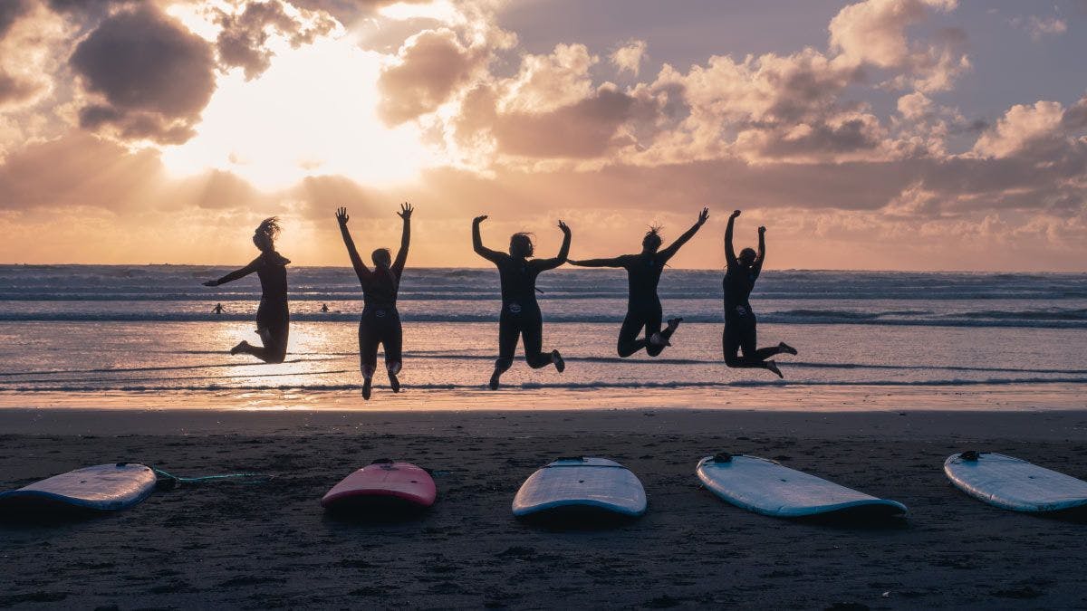 Group of surfing friends jump in the air on a beach in New Zealand