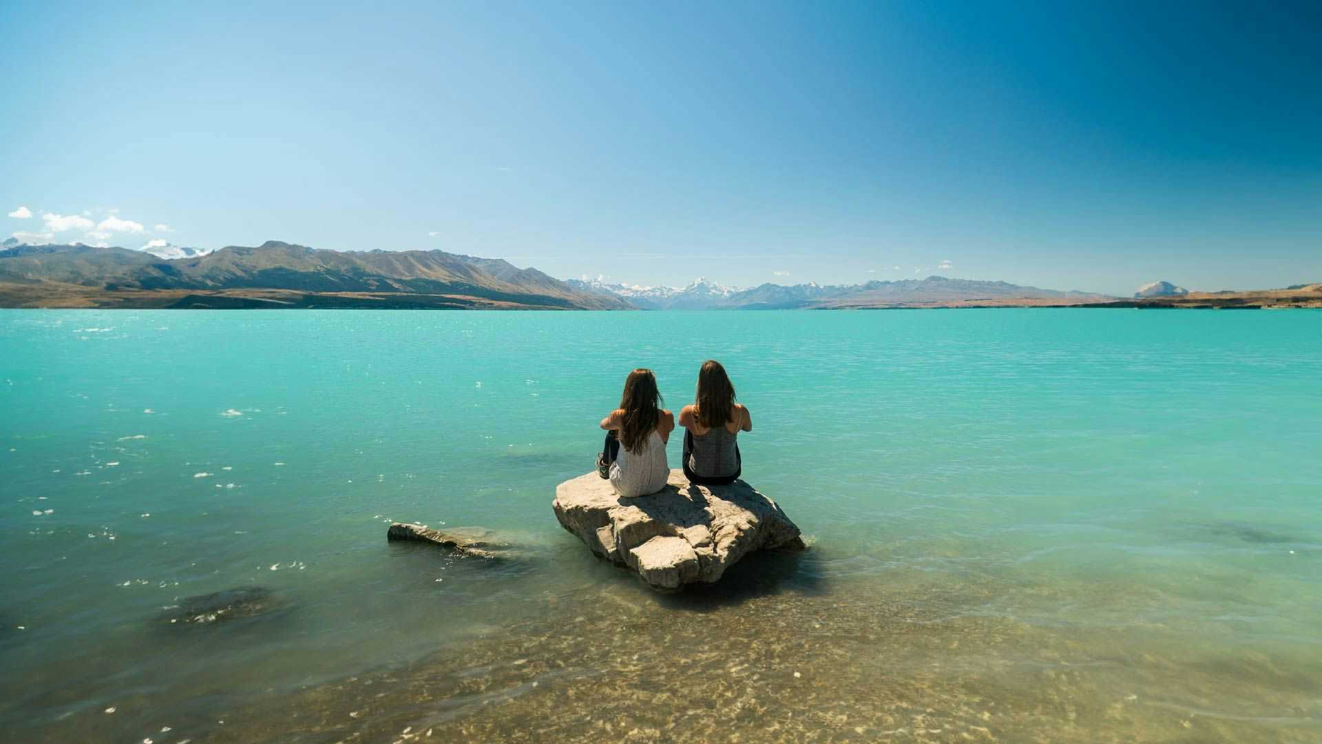 Two women sitting on a rock looking out at Lake Tekapo